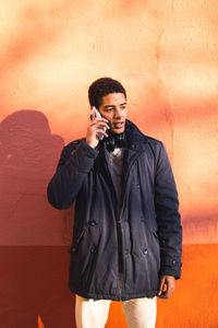 Young man using phone while standing against wall