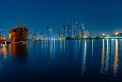 Illuminated buildings by river against blue sky at night