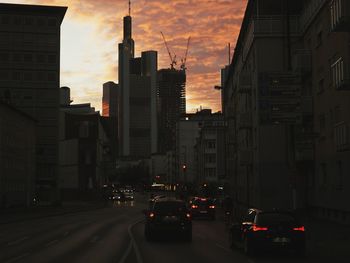 Cars on city street by buildings against sky during sunset