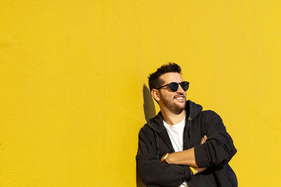 Young bearded man standing against a yellow wall with crossed arms