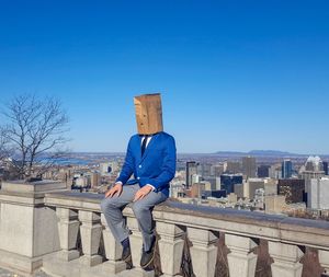 Man covered with paper bag sitting on railing against clear blue sky
