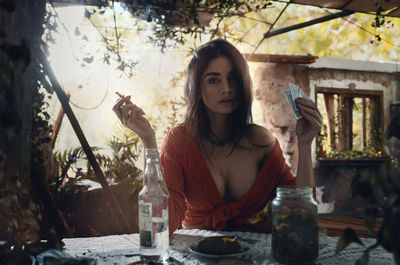 Portrait of beautiful woman holding playing card and smoking cigarette sitting by table in abandoned building