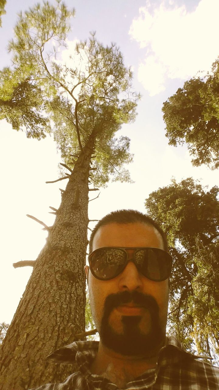 tree, low angle view, young adult, looking at camera, portrait, sky, front view, headshot, person, lifestyles, day, young men, leisure activity, built structure, outdoors, building exterior, branch, sunglasses