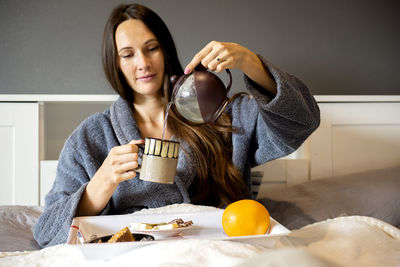  brunette woman sitting on the bed,holding tea cup and eating breakfast,morning routine concept