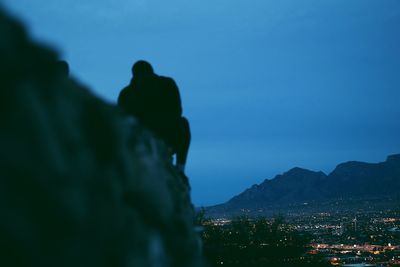 Silhouette man sitting on cliff against blue sky in city at dusk