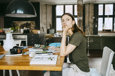 Female food stylist with hand on chin looking away while sitting at table in studio