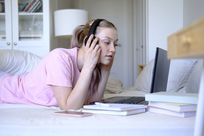 A young girl of 15-18 years old lies on a bed with a laptop and headphones and phone