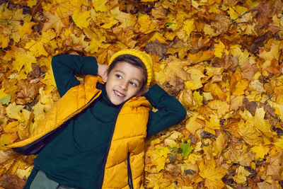 A boy child in a yellow jacket and hat lies on autumn leaves in the forest
