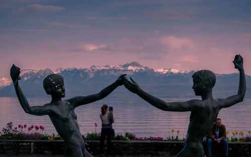 Statue standing by sculpture against sky during sunset