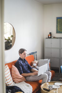 Man using laptop while sitting by toddler son drinking milk from bottle on sofa in apartment