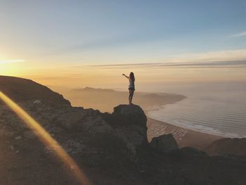 Man standing on rock by sea against sky during sunset