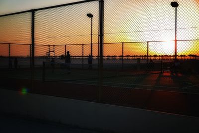 Silhouette people seen through chainlink fence against sky during sunset