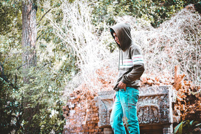 Side view of man wearing hood standing against trees in forest