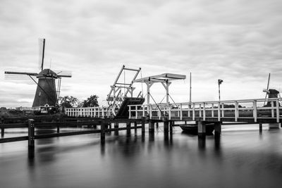 Windmill by pier over river against cloudy sky