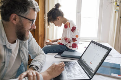 Smiling man working on laptop while daughter using smart phone at table