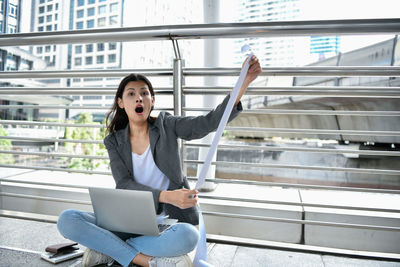 Portrait of shocked businesswoman with laptop holding bill on elevated walkway