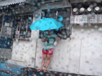 Woman with umbrella standing by building seen through wet window during monsoon