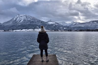 Rear view of mid adult woman in warm clothing standing on pier over lake against cloudy sky