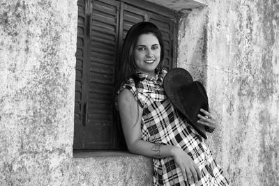 Portrait of woman holding hat while standing against wall