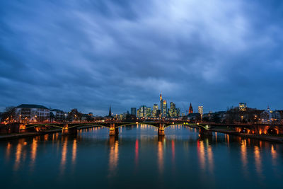 Illuminated bridge over river by buildings against sky at night