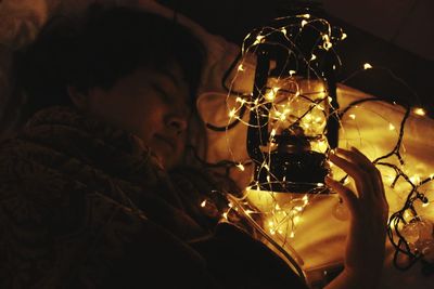 Close-up of woman with illuminated fairy lights and lantern in bedroom