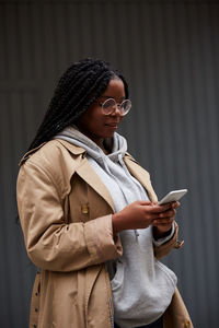 Young african american female in casual clothes and glasses standing on city street and browsing smartphone