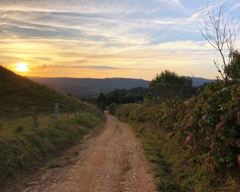 Dirt road passing through landscape against sky during sunset