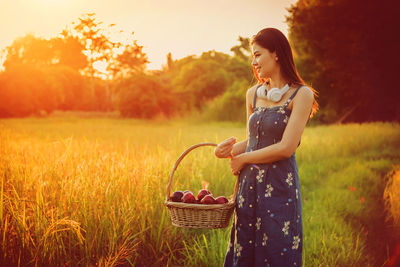 Smiling young woman carrying apples in wicker basket while standing on grassy field during sunset