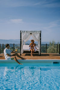 Couple sitting by swimming pool against sky