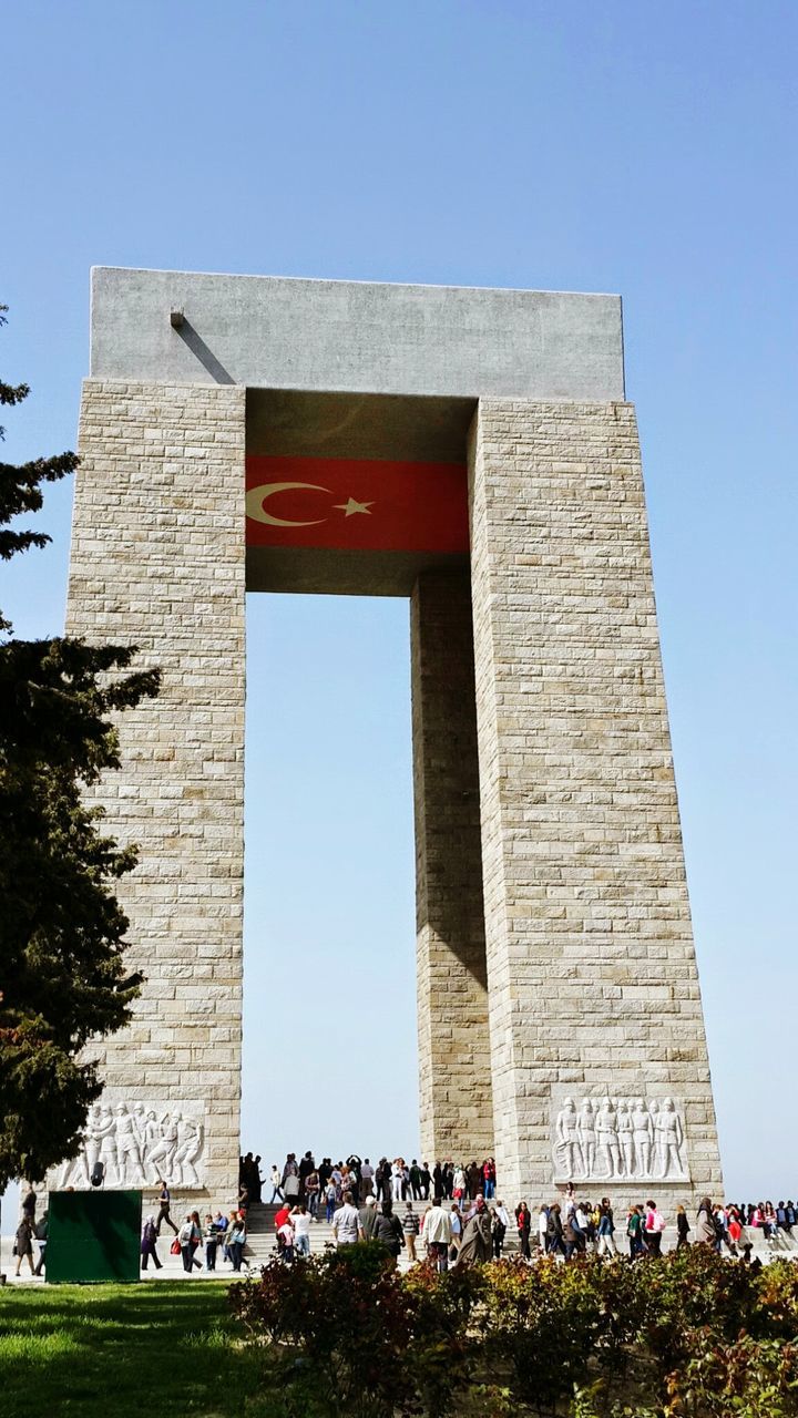 architecture, building exterior, built structure, clear sky, low angle view, blue, flag, tree, large group of people, day, copy space, sunlight, person, outdoors, tower, men, guidance, history, patriotism