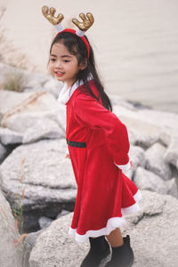 Side view of cute girl wearing red dress and headband standing on rocks at beach