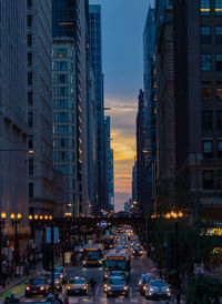 Traffic on city street by buildings against sky at dusk