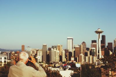 Rear view of man photographing space needle amidst buildings in city against clear sky on sunny day