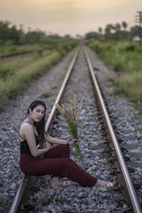 Woman holding plant while sitting on railroad track during sunset