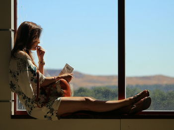 Full length of woman sitting on window sill against clear sky during sunny day
