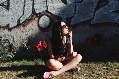 Thoughtful young woman sitting on field against graffiti wall