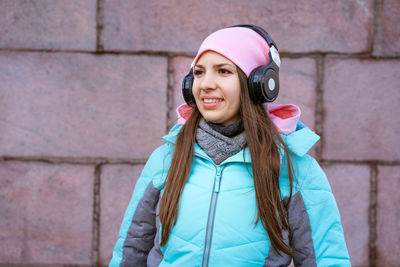 Happy young woman in warm jacket and pink hat listening to music on headphones