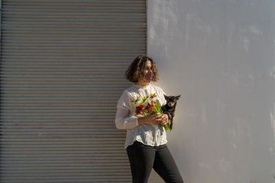 Young woman with mask holding bouquet of colorful flowers with her dog, white wall background