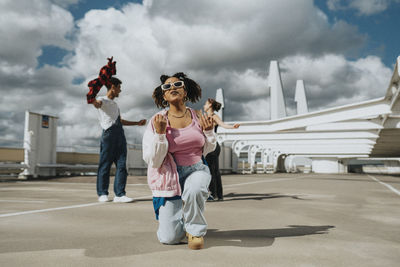 Carefree fashionable young woman kneeling and singing in parking lot under cloudy sky