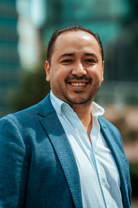 Happy hispanic mature male manager in suit holding hands in pockets and looking at camera with smile while walking near glass building on sunny day on city street