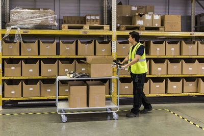 Full length side view of young male worker pushing cart with cardboard boxes by rack at distribution warehouse