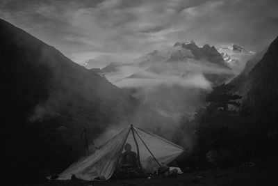 Man sitting in tent against mountains and foggy sky at night