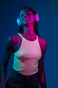 Positive brazilian female in stylish wear listening to music in wireless headphones while standing on blue background in studio with fluorescent light