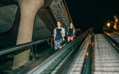 Women friends training running down escalator in city at night