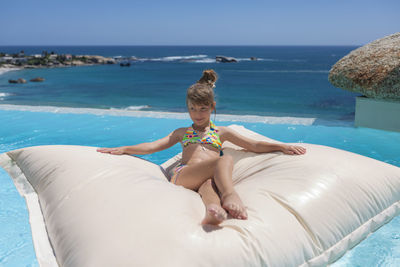 Girl wearing bikini lying on pool raft in infinity pool against clear blue sky