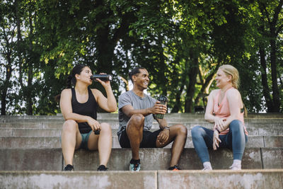 Low angle view of smiling male and female friends drinking water and talking during break after workout while sitting on
