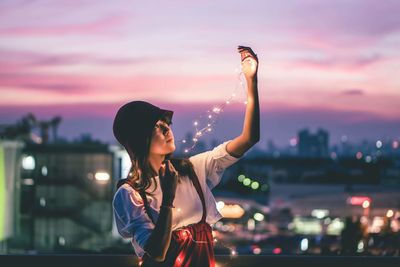 Young woman standing by illuminated city against sky during sunset