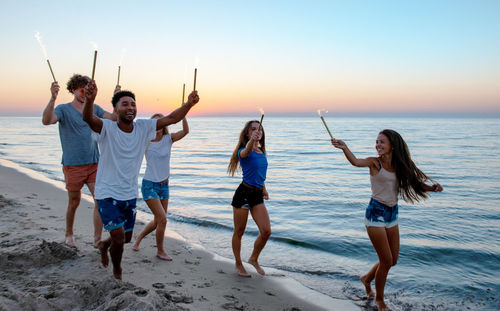 Cheerful friends holding sparkler walking at beach during sunset