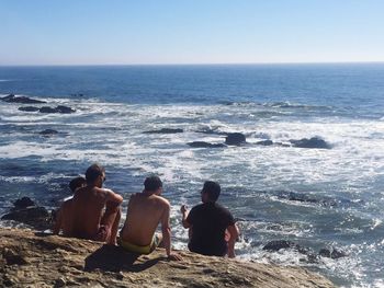 Rear view of men sitting on rock by sea against clear sky