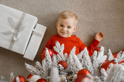 Blond little boy in red sweater wrinkles nose while laying under the decorated tree with presents